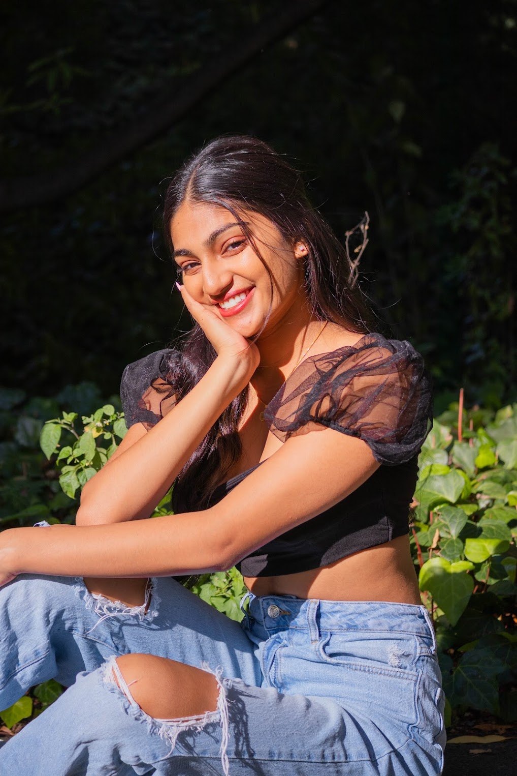 Mahika Valluri smiling in a garden, black tulle-sleeve top and ripped jeans