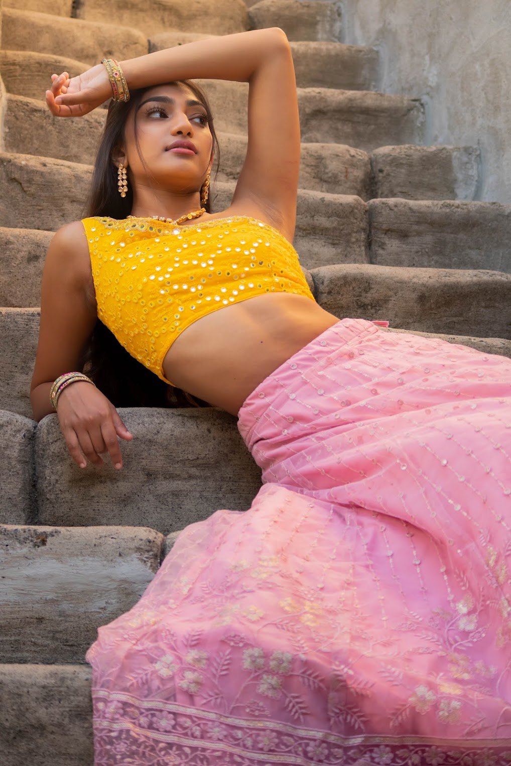 Mahika Valluri reclining on stone stairs in a yellow blouse and pink lehenga