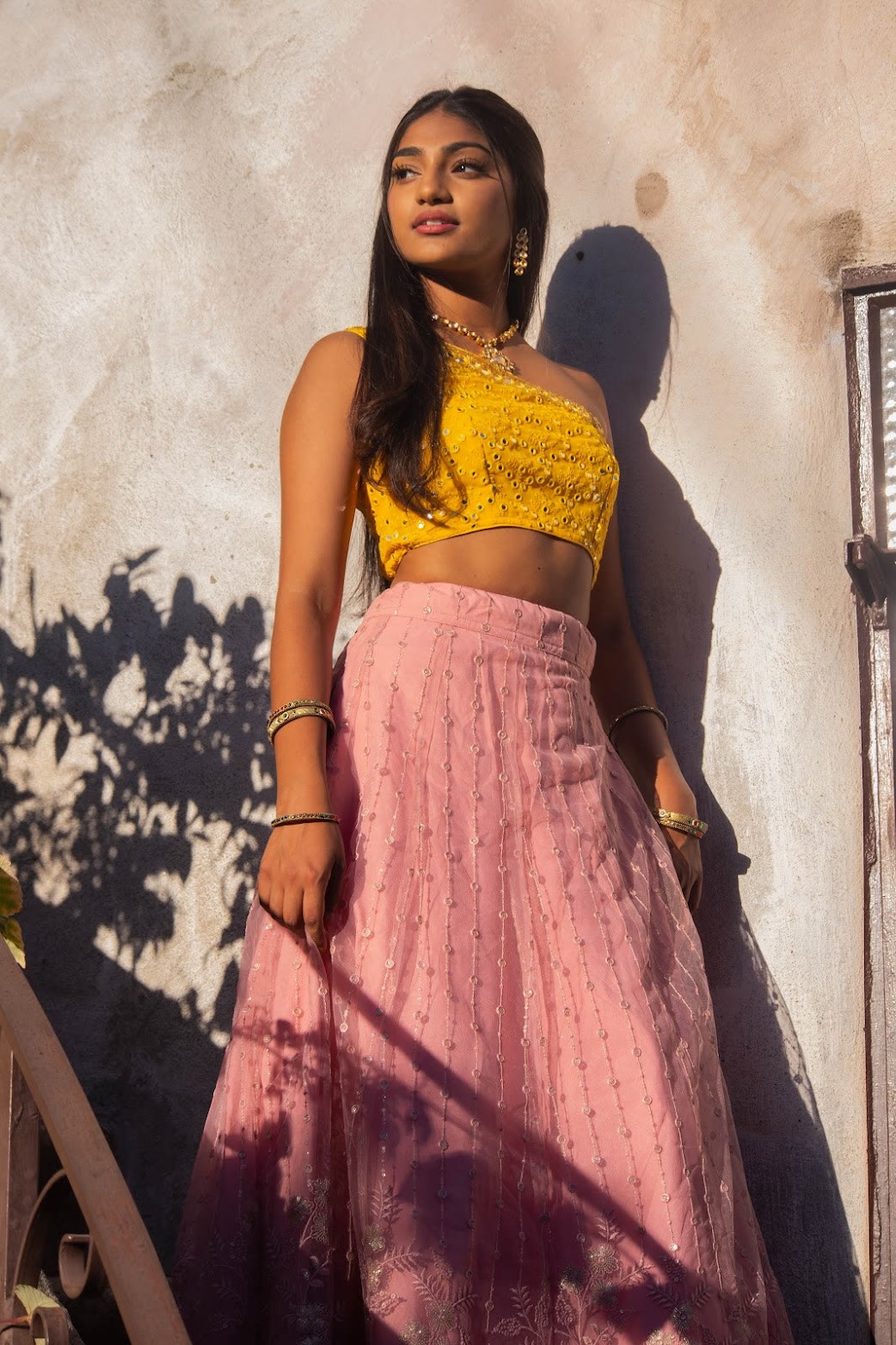 Mahika Valluri in a yellow mirror-work blouse and pink lehenga against a sunlit wall